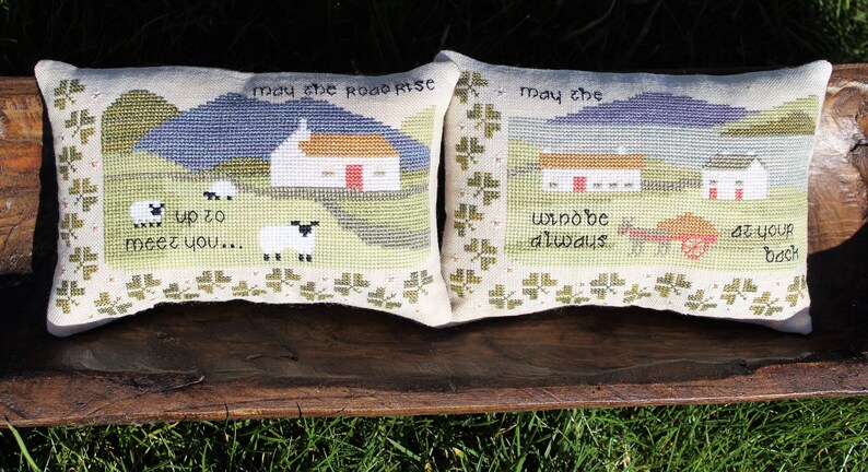 Puede incluir: Dos fundas de coj&iacute;n bordadas con una escena tradicional del campo irland&eacute;s. Las almohadas presentan una caba&ntilde;a blanca, colinas verdes y ovejas. El texto en las almohadas dice "Que el camino se eleve para encontrarte..." y "Que el viento est&eacute; siempre a tu espalda."