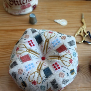 Op de afbeelding: Een wit stoffen naaldenkussen met een rond ontwerp met verschillende naai-accessoires, waaronder scharen, naalden en spoelen garen. Het ontwerp is in tinten rood, blauw en goud.