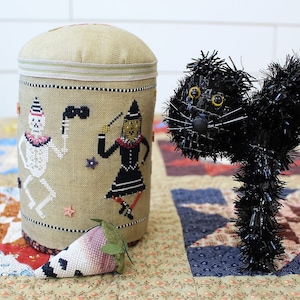 May include: A beige fabric cylinder with a black and white cross-stitch design of a skeleton and a witch. The cylinder is sitting on a patchwork quilt with a black cat figurine in the background.