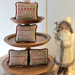 May include: A three-tiered wooden serving stand with four embroidered fabric squares. The squares are green with red embroidery and feature Christmas-themed text. The text on the squares reads "Mincepies Mulled Wine", "Christmas Crackers & Pudding", "Carols Candle Light", and "Holly Mistletoe Ivy". A white porcelain Santa Claus figurine is standing to the right of the stand.