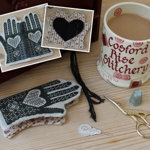 May include: A pair of black and white needlepoint hand-shaped pincushions with a heart design. The pincushions are on a wooden surface with a cup of tea, scissors, and a thimble.