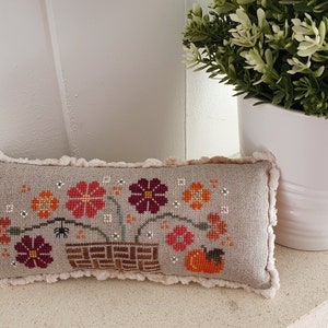 May include: A rectangular, beige cross-stitch pillow featuring a floral design in shades of red, orange, and burgundy, with a small spider and pumpkin. The pillow has a fluffy, cream-colored trim and is placed on a white surface.