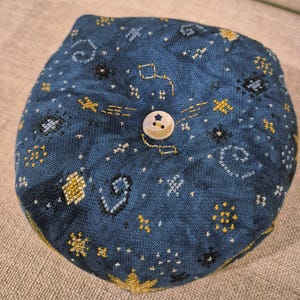 May include: A round, blue pincushion with a celestial design. The fabric is embroidered with gold, white, and black stars, constellations, and other shapes. A white button with a gold crescent moon and star detail secures the top.