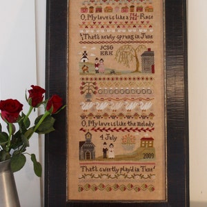 Könnte beinhalten: Ein gerahmter Kreuzstich-Sampler mit beigefarbenem Hintergrund und bunter Stickerei. Der Sampler zeigt ein Gedicht über die Liebe und eine Hochzeitsszene mit einer Kirche, einem Paar und dem Datum "4. Juli 2009".
