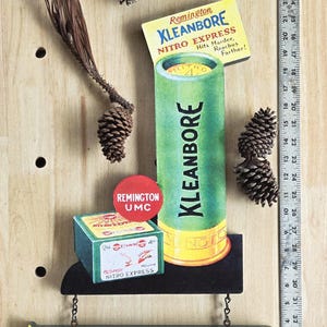 May include: Vintage Remington Kleanbore Nitro Express advertising display. The display features a large green and yellow shotgun shell, a box of shells, and a tall green Kleanbore shell. Pinecones and a ruler are also present.
