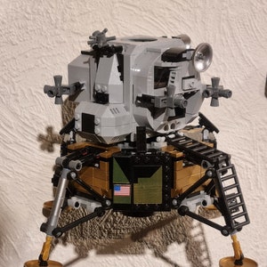 May include: A gray and gold Lego model of the lunar module, the spacecraft that landed on the moon. The model has a detailed interior and exterior, including a ladder, landing legs, and an American flag.