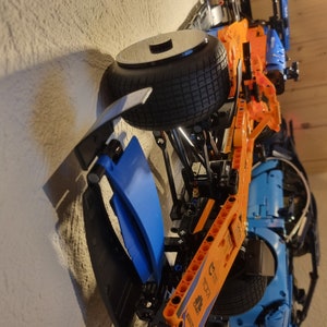 May include: A detailed model of a Formula 1 race car, featuring a black tire, orange and blue body panels, and visible mechanical components. The model is mounted on a wall, showcasing its design.