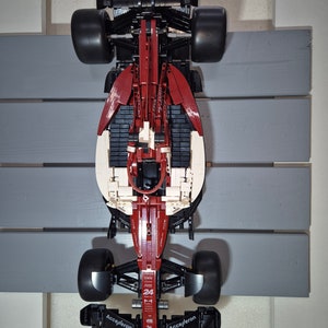 May include: A red and black Lego Formula 1 race car with the words "Accel/eron" and "MULD" on the front. The car is on a gray wooden surface.