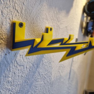 May include: Three yellow lightning bolt shaped wall hooks with blue accents. The hooks are mounted on a white wall.