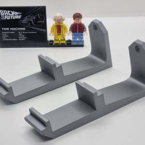 May include: Two gray 3D printed wall mounts for displaying Lego minifigures. The mounts have a curved shape and a small hole at the top for hanging.