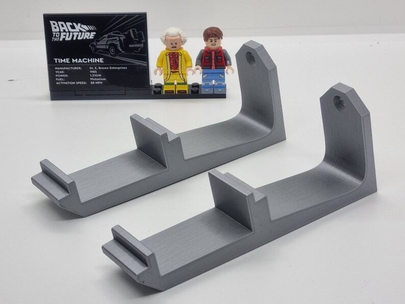 May include: Two gray 3D printed wall mounts for displaying Lego minifigures. The mounts have a curved shape and a small hole at the top for hanging.