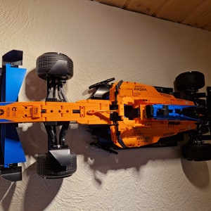 May include: An orange and blue Lego Technic McLaren Formula 1 race car model. The model features black tires, sponsor logos, and aerodynamic elements. The car is mounted on a wall.