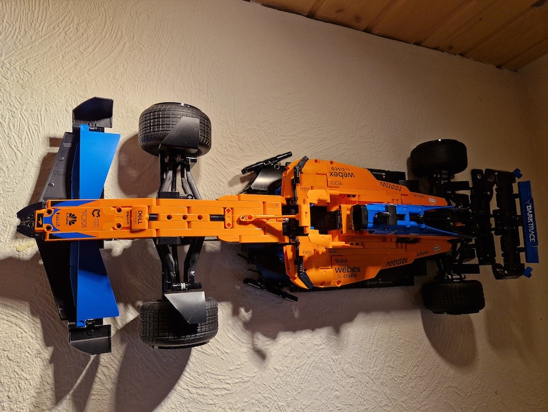 May include: An orange and blue Lego Technic McLaren Formula 1 race car model. The model features black tires, sponsor logos, and aerodynamic elements. The car is mounted on a wall.