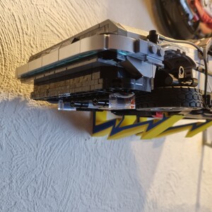 May include: A close-up of a gray and black Lego model of the DeLorean time machine from the Back to the Future movie. The model is mounted on a wall with yellow lightning bolt decals.
