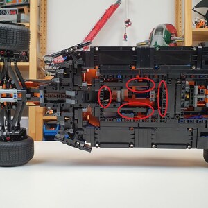 May include: A black and blue model car constructed from interlocking plastic bricks. The underside of the vehicle is visible, showcasing intricate mechanical details, including wheels, gears, and structural components. The model is set against a backdrop of shelves filled with other models.