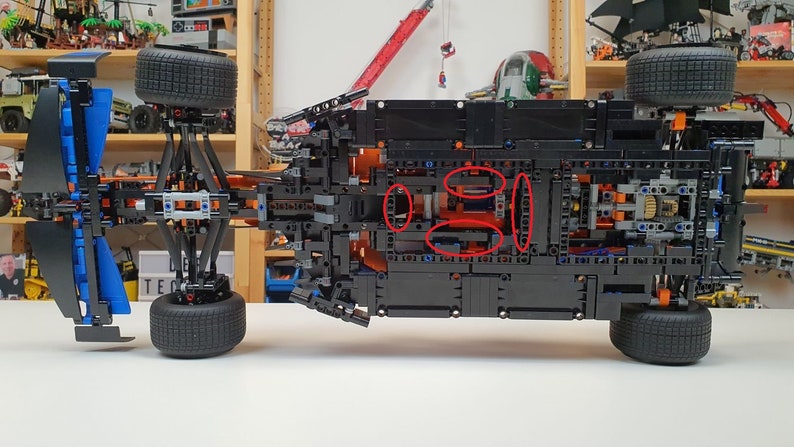 May include: A black and blue model car constructed from interlocking plastic bricks. The underside of the vehicle is visible, showcasing intricate mechanical details, including wheels, gears, and structural components. The model is set against a backdrop of shelves filled with other models.