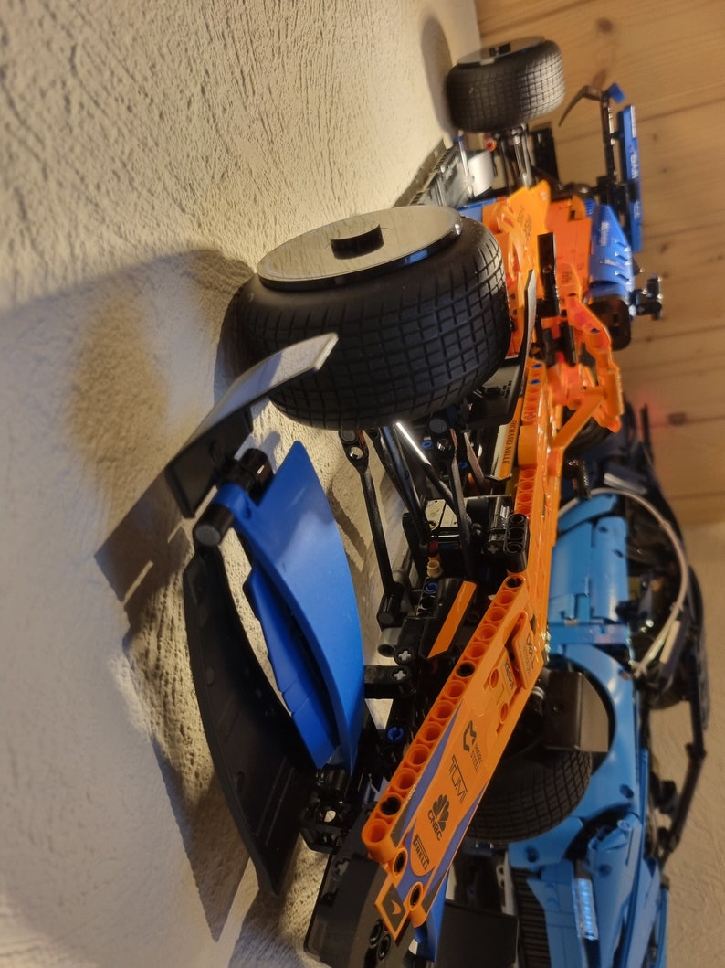 May include: A detailed model of a Formula 1 race car, featuring a black tire, orange and blue body panels, and visible mechanical components. The model is mounted on a wall, showcasing its design.