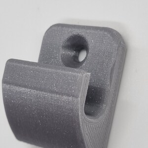May include: A gray, 3D-printed hook with a curved design. The hook features a rounded top with a hole for mounting and a curved lower section for hanging items. The surface has visible layer lines from the printing process.