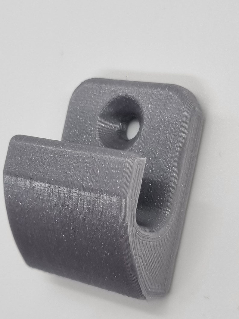May include: A gray, 3D-printed hook with a curved design. The hook features a rounded top with a hole for mounting and a curved lower section for hanging items. The surface has visible layer lines from the printing process.