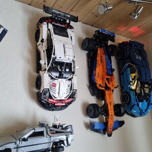 May include: A collection of model cars, including a white Porsche 911 with black accents and a blue McLaren, mounted on a wall. The Porsche has "PORSCHE" on the spoiler. A silver DeLorean model is also visible.