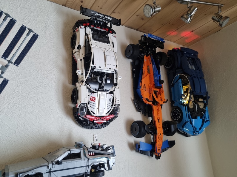 May include: A collection of model cars, including a white Porsche 911 with black accents and a blue McLaren, mounted on a wall. The Porsche has "PORSCHE" on the spoiler. A silver DeLorean model is also visible.