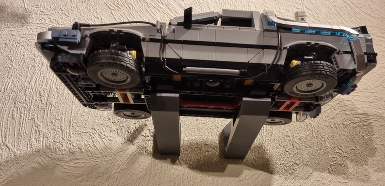 May include: A gray and black Lego model of the DeLorean time machine from the Back to the Future movie series. The model is mounted on a wall-mounted stand.