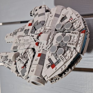 Könnte beinhalten: Ein detailliertes Modell des Raumschiffs Millennium Falcon, aus LEGO-Steinen gebaut. Das Modell ist grau und weiß mit roten Akzenten. Das Schiff hat ein Cockpit, Flügel und einen Heckmotor.