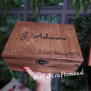 May include: A wooden box with a hinged lid and a metal clasp. The box is stained a dark brown colour and has the text "J. Ashmore 22nd February 1979" engraved on the top. The box is being held by a hand with red nail polish.