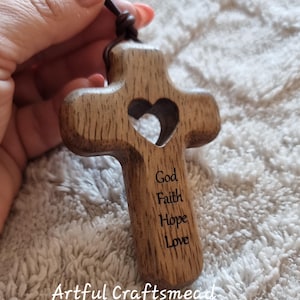 Könnte beinhalten: Holz-Kreuz-Anhänger mit herzförmigem Ausschnitt und den eingravierten Worten "Gott, Glaube, Hoffnung, Liebe". Es hängt an einer braunen Lederschnur. Der Text "Artful Craftsmead" befindet sich unten.