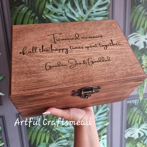 Könnte beinhalten: Eine Holz-Andenkenbox mit Scharnierdeckel und Metallverschluss. Die Box ist mit den Worten "Treasured memories of all the happy times spent together... Grandma, Jen & Granddad" graviert. Die Box wird in einer Hand gehalten.