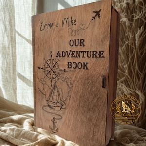 以下が含まれることがあります： コンパスローズ、ヨット、錨、飛行機、そして「Emma & Mike OUR ADVENTURE BOOK」という文字がレーザー刻印された、蝶番付きの蓋を持つ長方形の木製ボックス。木材は自然な木目調の仕上げが施されています。