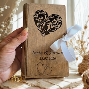 May include: A rectangular wooden box with a laser-cut filigree heart design on the front. The box features the engraved text Ineta & Aarian and the date 25.07.2026, with two small line-art hearts below. A white ribbon is tied around the side.