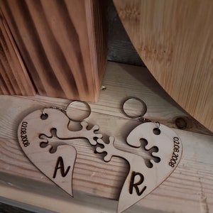 May include: Two wooden heart-shaped keychains with a puzzle-like design that fit together. The keychains are engraved with the letters "N" and "R" and the date "02082008".