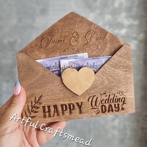 Op de afbeelding: Een houten envelop met een hartvormige uitsparing en de tekst "Happy Wedding Day" erop gegraveerd. De envelop is open en bevat contant geld. De envelop is gepersonaliseerd met de namen "Jaime & Paul" en de datum "13-07-2024".