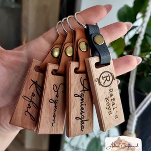 Könnte beinhalten: Eine Sammlung personalisierter Holzschlüsselanhänger. Jeder Schlüsselanhänger hat ein rechteckiges Holzschild mit einem handschriftlich eingravierten Namen. Die Schlüsselanhänger sind an Lederriemen mit Metallnieten und Schlüsselringen befestigt. Ein Schlüsselanhänger trägt den Text "Rafal's keys".