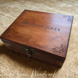 May include: A dark brown wooden box with the name "Sebastian Jukes" engraved on the lid. The box has decorative corner details and a metal clasp. The box is likely made of wood.