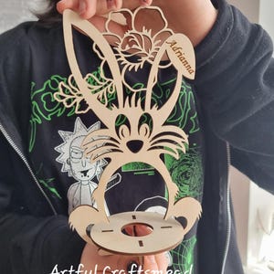 May include: Wooden Easter bunny egg holder with a floral design and the name "Adriana" engraved on the side. The holder is made of light brown wood and has a round base.