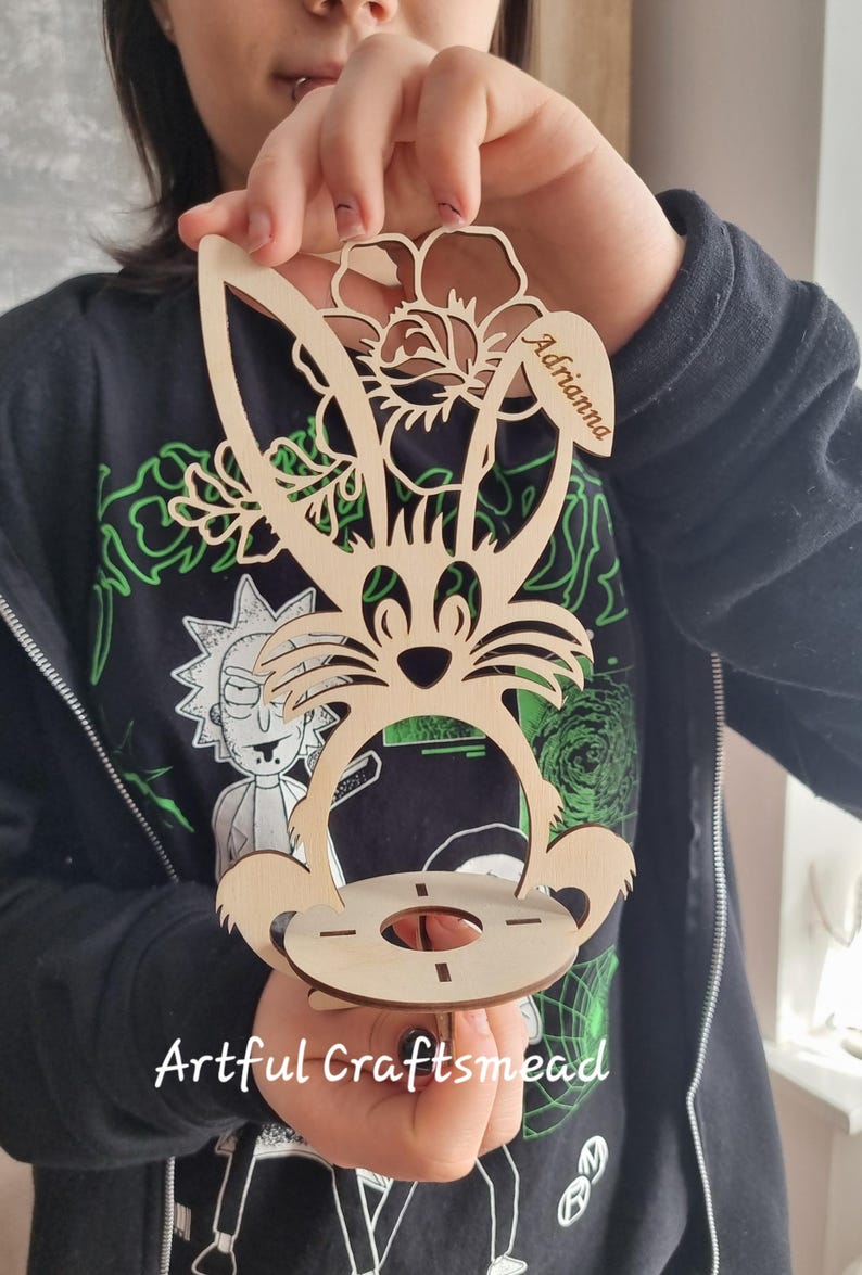 May include: Wooden Easter bunny egg holder with a floral design and the name "Adriana" engraved on the side. The holder is made of light brown wood and has a round base.