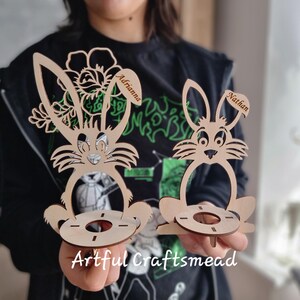 May include: Two wooden bunny-shaped egg holders with personalized names "Adriana" and "Nathan". The holders are made of light brown wood and have a cutout design of a bunny with a flower on the top. The holders are sitting on a table.