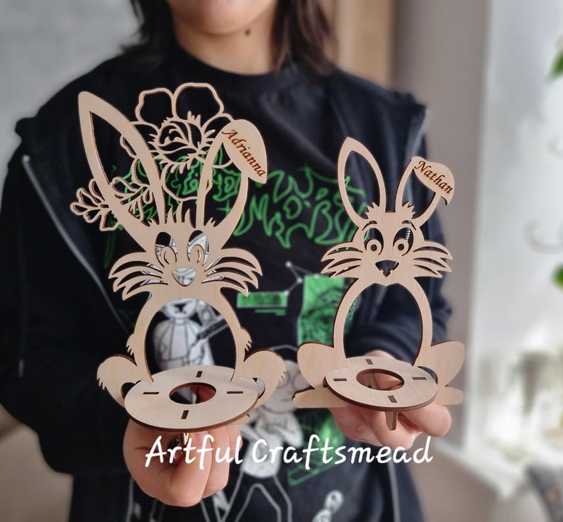 May include: Two wooden bunny-shaped egg holders with personalized names "Adriana" and "Nathan". The holders are made of light brown wood and have a cutout design of a bunny with a flower on the top. The holders are sitting on a table.