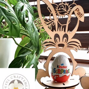 May include: Wooden Easter bunny stand with a flower design and the name "Pati" engraved on the side. A Kinder Surprise egg is placed in the stand.