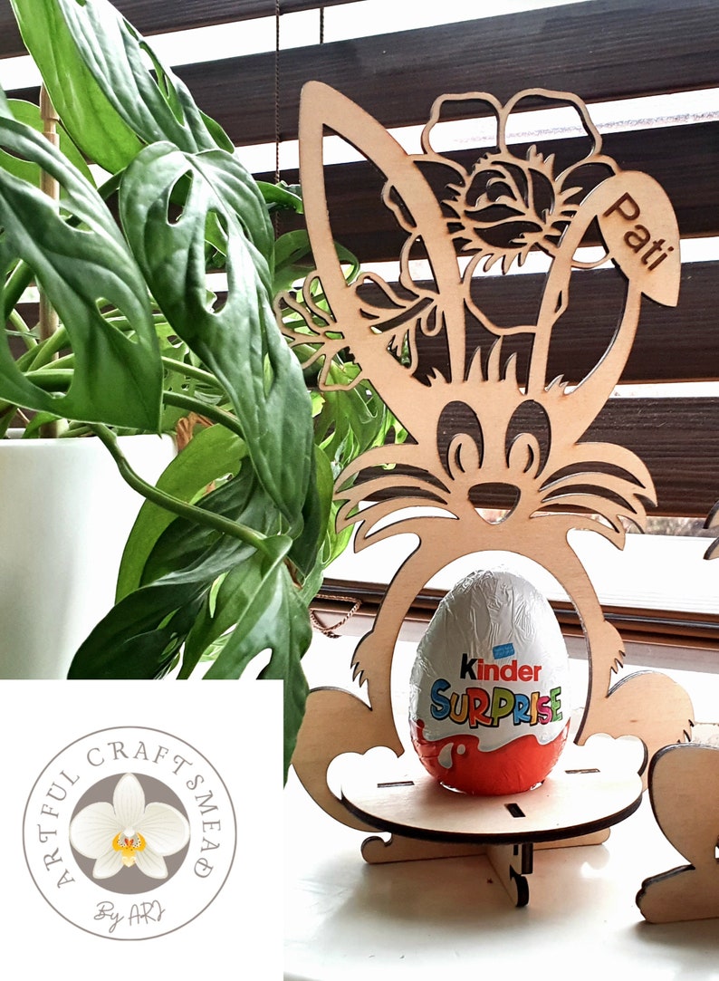 May include: Wooden Easter bunny stand with a flower design and the name "Pati" engraved on the side. A Kinder Surprise egg is placed in the stand.