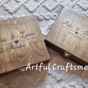 May include: Two wooden boxes with engraved names and dates. One box reads "Chris & Lauren 21st November 2025" with a heart symbol. The other box reads "Risha & Dillon 07.11.2025" with a heart symbol. The boxes are light brown.