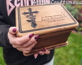 May include: A wooden box with a cross and floral design engraved on the lid. The lid is inscribed with "in loving memory of KATE HOLLOWAY 01/05/1963-12/01/2023".