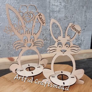 May include: Two wooden bunny egg holders with names "Kasia" and "Stawek" written on them. The bunny holders are sitting on a wooden surface. The bunny holders are a craft project from Artful Craftsmead.