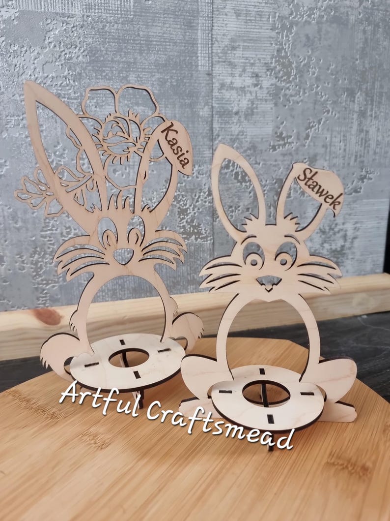 May include: Two wooden bunny egg holders with names "Kasia" and "Stawek" written on them. The bunny holders are sitting on a wooden surface. The bunny holders are a craft project from Artful Craftsmead.