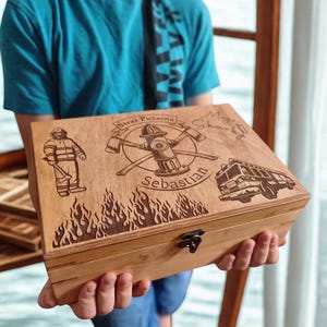 May include: A wooden box with a hinged lid, featuring engraved firefighter-themed designs. The lid displays a firefighter, fire truck, and a fire hydrant with crossed axes. The name "Sebastian" is also engraved. Flames are etched along the bottom edge.