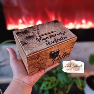 May include: A wooden money box with a floral design and the text "Moja pierwsza skarbonka" engraved on the front. The money box has a slot for coins and a metal latch.