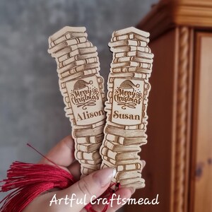 May include: Two wooden Christmas bookmarks shaped like stacks of books. Each bookmark has the words "Merry Christmas" and a name, "Alison" and "Susan", engraved on them. A red tassel hangs from the bottom of the bookmarks. The text "Artful Craftsmead" is also visible.