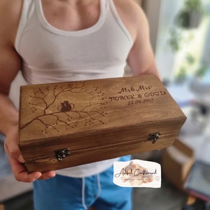 May include: A dark brown wooden box with a laser-etched tree design, two birds, and the inscription "Mr & Mrs TOMEK & GOSIA 25.06.2012". The box is secured with metal clasps and hinges.
